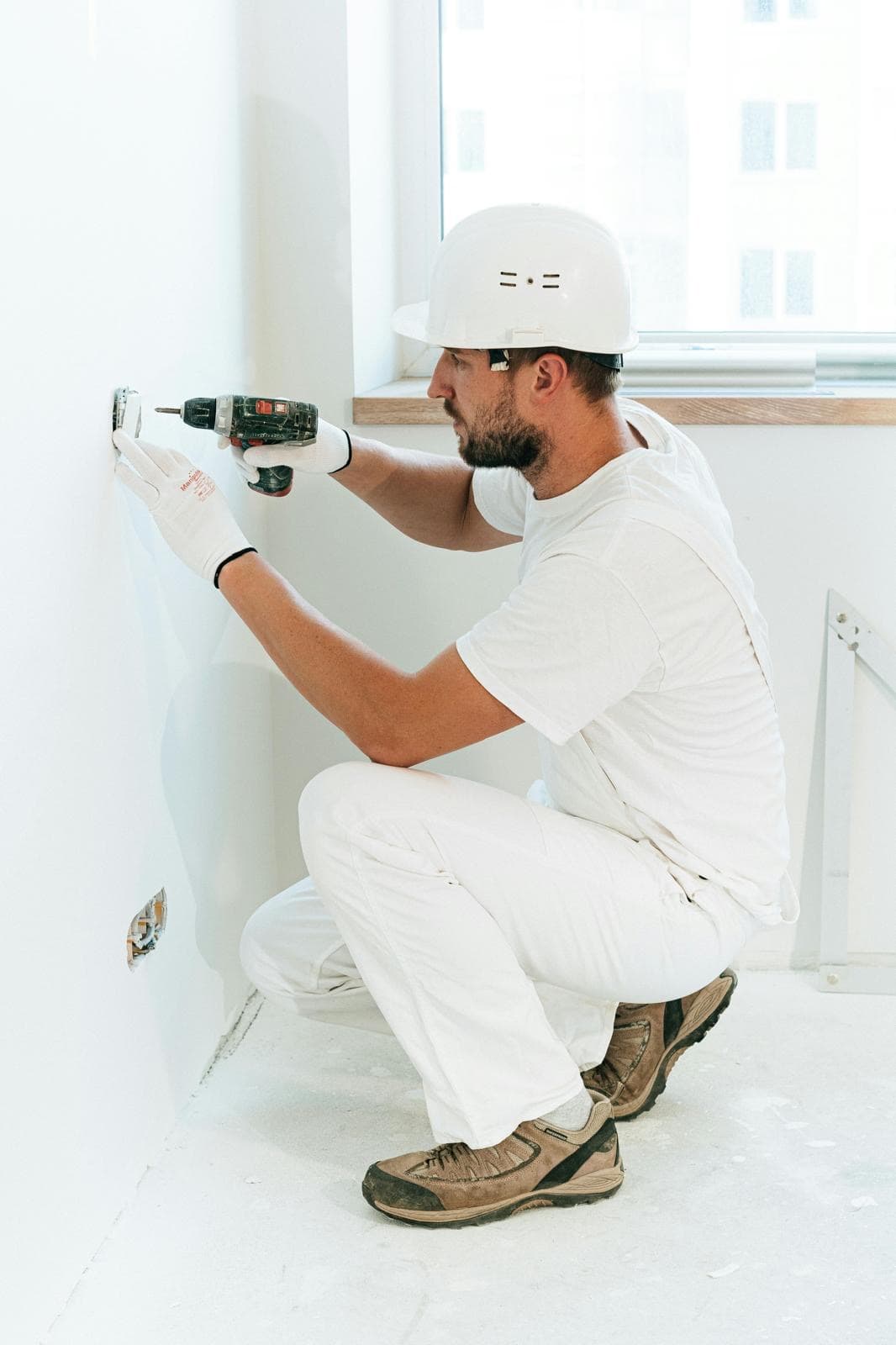 Worker in white hard hat and clothing uses a drill on a wall outlet.